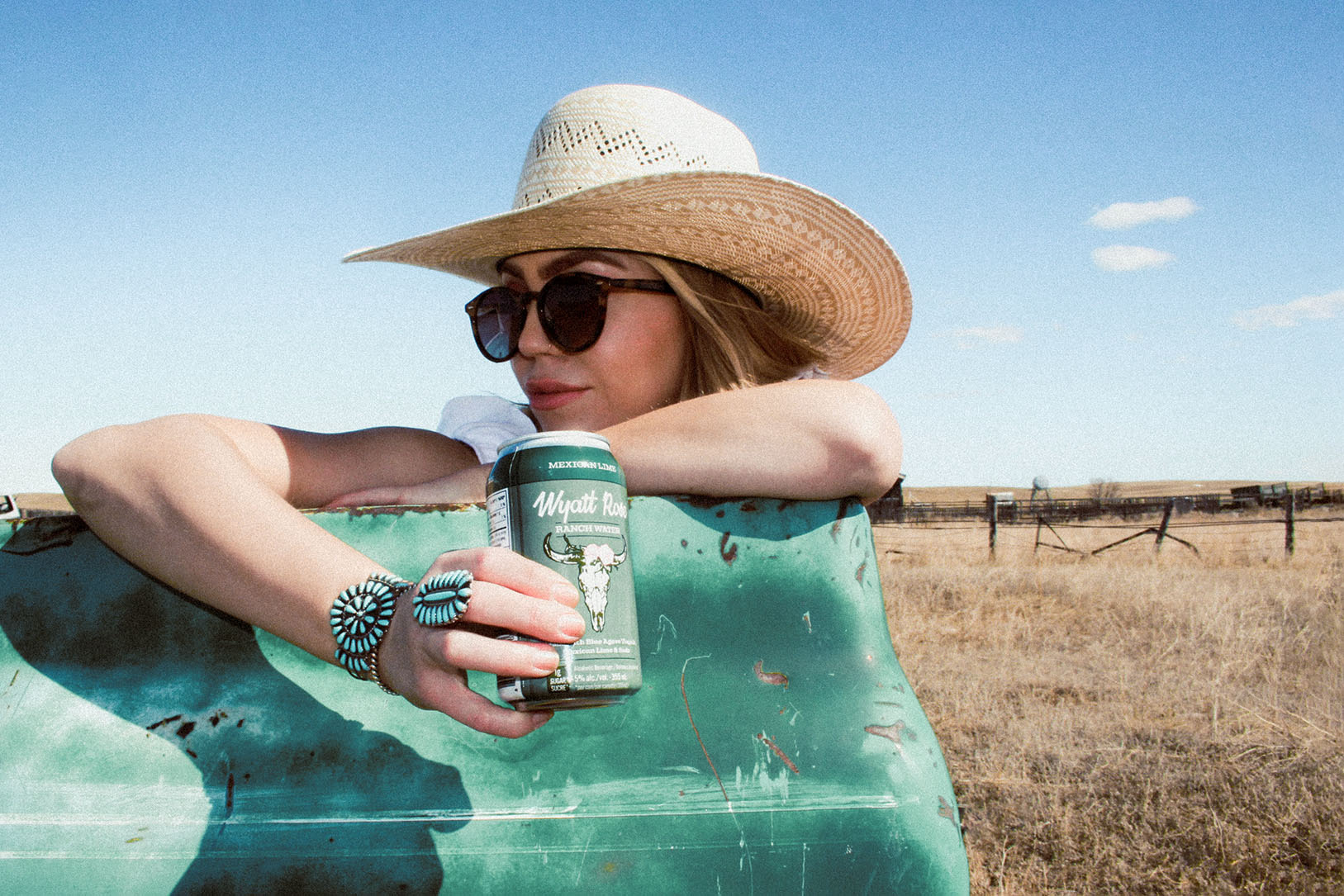 Woman in cowboy hat leaning over a rusty truck door, holds a can of Wyatt Rose Ranch Water in a field in the prairies