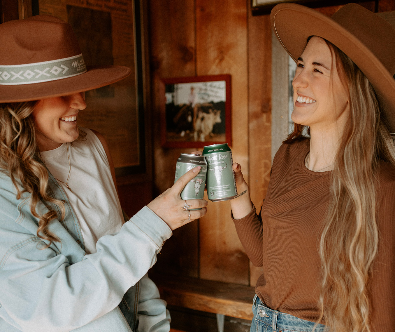 Two women in cowboy hats cheers with a can each of Wyatt Rose Ranch Water